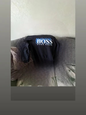 Svart sweatshirt från Hugo Boss - Snygg svart sweatshirt från Hugo Boss med stort vitt och blått BOSS-tryck på bröstet. Tröjan har rund halsringning med blå kant och långa ärmar. Perfekt för dig som vill ha en stilren och trendig look.