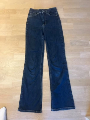 Mörkblå bootcut jeans - Snygga mörkblå bootcut jeans med hög midja och klassiska fem fickor. Jeansen har en rak passform upptill och utsvängda ben, vilket ger en retrovibe. Tillverkade i denim med kontrastsömmar och knappgylf. 