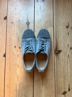 Loubs - Säljer ett par gråa sneakers från Louboutin med ikoniska röda sulor och coola nitar på tårna. Skorna har snörning och är tillverkade i mocka för en lyxig känsla. Perfekta för dig som vill sticka ut med din stil. Hör av dig vid frågor🙌🏼