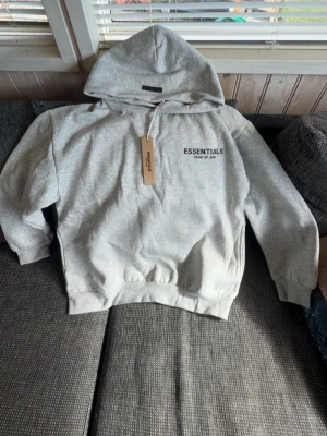 Essentials hoodie - Snygg grå hoodie från Essentials Fear of God med svart logga tryckt på bröstet och ryggen. Klassisk passform med huva och ribbade muddar vid ärmar och midja. Perfekt för en avslappnad streetwear-look. Tillverkad i mjukt sweatshirtmaterial.