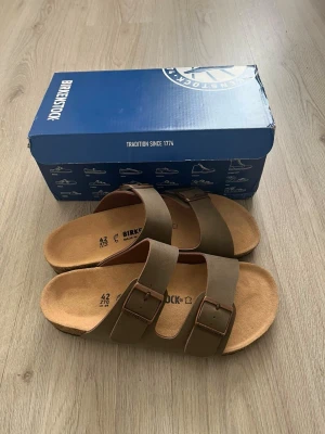 Bruna Birkenstock sandaler med spännen - Säljer ett par klassiska bruna Birkenstock sandaler med två breda remmar och justerbara metallspännen. Fotbädden är i mocka och sulan är ergonomiskt utformad för extra komfort. Perfekta för en avslappnad och stilren look.