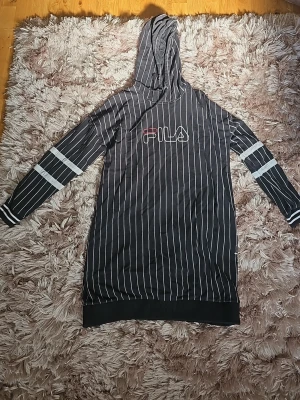 Svart randig hoodieklänning FILA - Säljer en svart hoodieklänning från FILA med vita tunna ränder och logga på bröstet. Långa ärmar med vita detaljer och ribbade muddar. Skön huva och avslappnad passform, perfekt för en chill och sportig look.