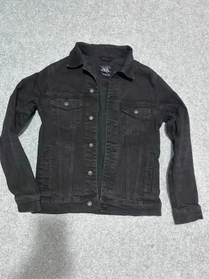 Svart jeansjacka från Zara - Snygg svart jeansjacka från Zara med klassisk krage och knäppning framtill. Jackan har två bröstfickor med lock och knapp samt två sidofickor. Tillverkad i slitstarkt denimtyg och har långa ärmar med knapp vid ärmslut. Perfekt för en cool och avslappnad stil.