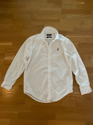 Vit skjorta från Ralph Lauren - Klassisk vit skjorta från Ralph Lauren med broderad logga på bröstet. Skjortan har lång ärm, knappar framtill och en tidlös passform. Perfekt för en clean och stilren look. Storlek 16 men passar som S.