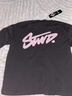 Svart t-shirt med rosa SWD-tryck - Säljer en svart t-shirt med stort rosa SWD-tryck på ryggen och mindre logga på bröstet. T-shirten har rund hals och korta ärmar. Perfekt för dig som gillar streetwear och enkel stil. Aldrig använt!!
