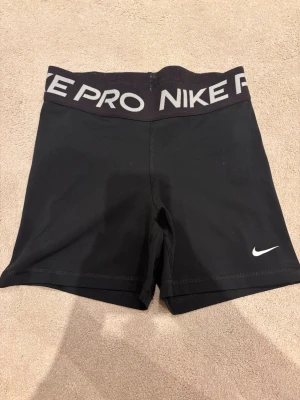 Svarta Nike Pro Dri-FIT shorts - Säljer ett par svarta Nike Pro Dri-FIT shorts med bred resår i midjan där det står 'NIKE PRO'. Materialet är stretchigt och andas, perfekt för träning eller gym. Klassisk vit Nike-logga på benet och tight passform.