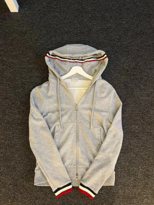 Grå Moncler zip hoodie  - Snygg grå hoodie med dragkedja framtill och huva med snörning. Röd, vit och svart rand på huva och ärmslut ger en sportig vibe. Perfekt till chill eller streetwear. Mjuk och skön bomullsblandning, två fickor fram.