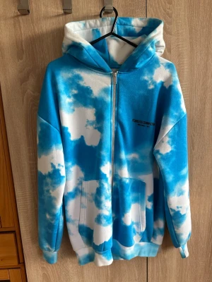 FIN TRÖJA - Säljer en oversized hoodie med blåvitt molnmönster och dragkedja framtill. Tryck på bröstet och ryggen, stor huva och rymliga fickor. Perfekt för dig som gillar streetwear och vill sticka ut med en unik look.