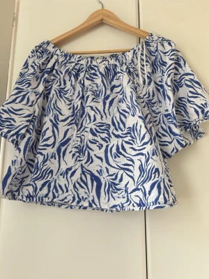 Blåvit offshoulder blus med zebramönster - Säljer en luftig offshoulder blus i blått och vitt med zebramönster. Toppen har korta, vida ärmar och resår vid halsen för en avslappnad look. Perfekt för varma dagar och ger en trendig vibe till din outfit.