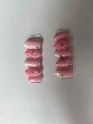 Rosa press-on naglar med guld och prickar - Säljer ett set press-on naglar i ljusrosa med vita prickar och små gulddetaljer. Naglarna har en kort, rundad form och en glansig finish. Perfekt för dig som vill ha en söt och trendig look utan att behöva gå till salong.