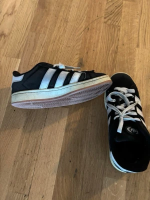 Svarta Adidas sneakers med vita ränder - Klassiska svarta sneakers från Adidas med de ikoniska tre vita ränderna på sidorna och vit sula. Skorna har vita skosnören och låg profil. Perfekta för en sportig och avslappnad stil. Pris kan diskuteras vid snabb affär!