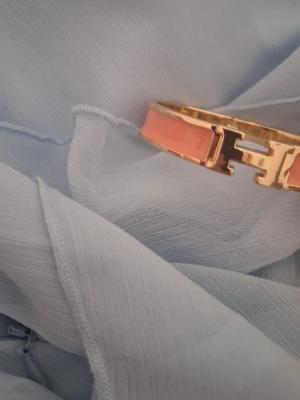 Guldigt armband - Snyggt armband i guldton med rosa emaljdetaljer och ett format spänne framtill. Armbandet har en stilren och modern design som sticker ut och passar perfekt till dig som gillar accessoarer med färg och metalliska inslag.