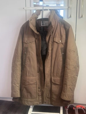 Brun rock med fickor och hög krage från diesel - Brun parkas med hög krage och fyra stora fickor framtill. Jackan har både dragkedja och knappar, samt detaljer i samma färg. Längre modell som ger extra skydd mot kyla. Perfekt för höst och vinter.