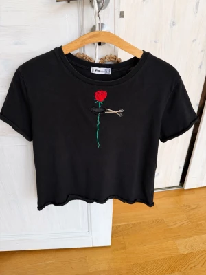 Svart crop top med rosbroderi - Svart crop top från FB Sister med korta ärmar och broderad röd ros samt sax på bröstet. T-shirten har rund halsringning och vågig nederkant. Materialet är mjukt och bekvämt, perfekt för en trendig och avslappnad stil.