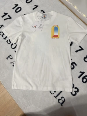Vit Casa Blanca t-shirt med tryck - Säljer en vit t-shirt från Casa Blanca med färgglatt tryck fram och stort grafiskt tryck på ryggen i gult, blått och rött. T-shirten har rund hals och korta ärmar. Perfekt för dig som gillar stilrena plagg med en twist.