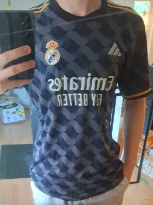 Real Madrid bortatröja Modric 10 - Snygg Real Madrid bortatröja från Adidas med Modric och nummer 10 på ryggen. Tröjan är mörkblå med guldiga detaljer, klubbmärke och Adidas-logga på bröstet samt sponsortryck. Tillverkad i lätt och ventilerande material, perfekt för fotboll eller träning.