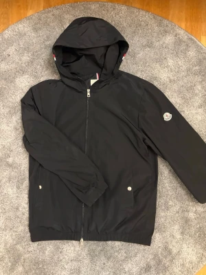 Svart vindjacka från Moncler - Snygg svart vindjacka från Moncler med huva och dragkedja framtill. Jackan har elastiska muddar och en diskret Moncler-logga på insidan. Huvan har en cool detalj med röd, vit och blå rand. Perfekt för blåsiga dagar.  Passar  M
