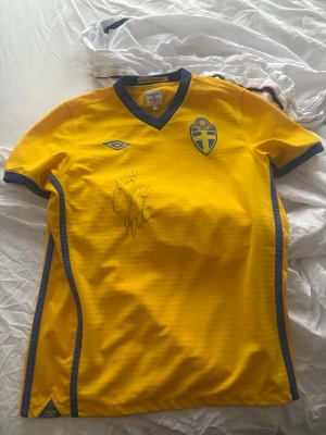 Svensk landslagströja Umbro gul med Zlatans autograf  - Gul svensk landslagströja från Umbro med blå detaljer och broderat landslagsmärke på bröstet. Tröjan har V-ringning och är signerad på framsidan. Perfekt för dig som älskar fotboll och vill sticka ut på läktaren. Är signerad av Zlatan