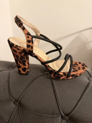 Leopardmönstrade sandaler med klack - Säljer ett par sandaler med chunky klack och leopardmönster. Skorna har flera tunna svarta remmar över foten och öppen tå. Klacken är hög och bred för extra stabilitet. Perfekta för dig som vill sticka ut med ett djurmönster.