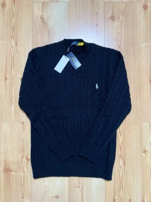 Svart kabelstickad tröja Polo Ralph Lauren - Kabelstickad svart tröja från Polo Ralph Lauren med rund halsringning och den klassiska broderade logotypen på bröstet. Tröjan har långa ärmar och ribbade muddar vid ärmslut och nederkant. Perfekt för dig som gillar stilren och tidlös design.