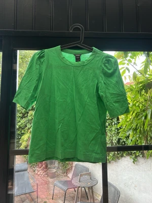 Grön topp med puffärm från Lindex - Snygg grön topp från Lindex i mjuk bomull. Modellen har puffärmar och rund halsringning. Perfekt att styla med jeans eller kjol för en fräsch outfit.