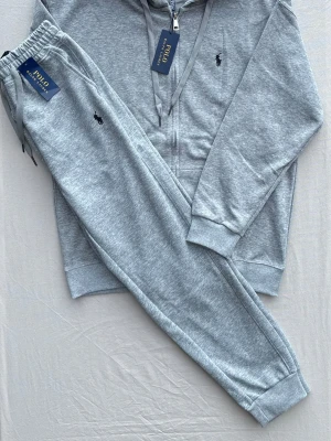 Grått loungewear-set från Polo Ralph Lauren - Mysigt grått loungewear-set från Polo Ralph Lauren med dragkedjetröja och matchande byxor. Setet har den klassiska broderade loggan på både tröjan och byxan. Tillverkat i mjuk bomullsblandning med ribbade muddar och justerbar huva.