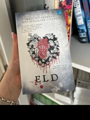 Eld av Mats Strandberg & Sara B. Elfgren - En spännande skönlitterär bok med titeln 'Eld' av Mats Strandberg och Sara Bergmark Elfgren. Omslaget är gråvitt med ett rött hjärta omgivet av svarta och vita rosor, och blodstänk. Perfekt för dig som gillar fantasy och mysterier.