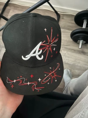 new era fitted Svart keps med broderade detaljer - Snygg svart keps med vit broderad A-logga framtill och röda samt vita stjärn- och swirlmönster på både skärm och baksida. Klassisk rak skärm och justerbar passform. Materialet är bomull och kepsen har en modern streetwear-vibe.