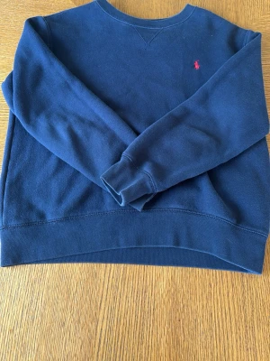 Mörkblå sweatshirt från Polo Ralph Lauren - Mörkblå sweatshirt från Polo Ralph Lauren med klassisk rund halsringning och liten röd logga broderad på bröstet. Tillverkad i mjukt och bekvämt material, perfekt för en avslappnad stil. Långa ärmar och ribbade muddar vid ärmslut och nederkant. (Lite bred passform)