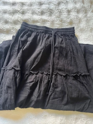 Svart långkjol med volang och snörning - Svart långkjol med volangdetaljer och snörning i midjan. Kjolen har en avslappnad passform och är tillverkad i ett mjukt tyg som känns skönt mot huden. Perfekt för dig som gillar en enkel men ändå trendig stil. Säljer därför jag tycker inte den passar mig längre. Jag är 160cm lång och den passar mig perfekt i längden! Knappt använd.