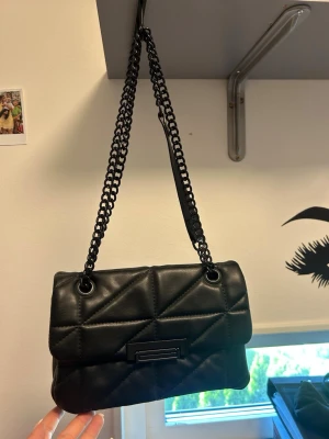 Svart quiltad handväska med kedja - Snygg svart handväska i quiltad design med rektangulär form och kedjerem. Väskan har ett lock med dekorativt spänne framtill och sydda geometriska mönster. Perfekt accessoar för att lyfta din outfit. Bandet går att ha både kort och långt