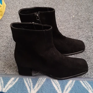 Svarta mockaboots från Russell & Bromley - Snygga svarta boots i mjuk mocka från Russell & Bromley, tillverkade i Italien. Modellen har en fyrkantig tå, låg klack och dragkedja på sidan för enkel på- och avtagning. Perfekt för dig som gillar stilrena och tidlösa skor med en modern twist.