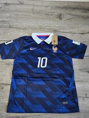 Frankrike fotbollströja Mbappe 2026 - Snygg blå fotbollströja från Nike med Frankrikes landslagsemblem och nummer 10 på bröstet. Tröjan har vit krage, korta ärmar och Mbappé samt nummer 10 på ryggen. Mönstret är diagonalrandigt i olika blå nyanser och materialet är lätt och andas.