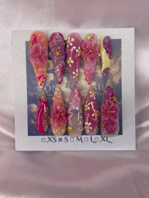 LuxeFantasy (S);ExtraLongStiletto - Set med unika och handgjorda/handmålade extra långa, stiletto press-on naglar i flera färger såsom rosa, lila och nude toner med handgjorda 3D-blommor, gulddekorationer och små vita handmålade blommor. Naglarna har en glansig finish och detaljer som stjärnor och pärlor för en extra lyxig look. Perfekt för dig som vill sticka ut med din nageldesign!S: 15 mm, 11,5 mm, 12 mm, 11 mm, 9 mm 