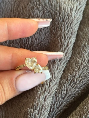 Guldfärgad ring med hjärtformade stenar - Snygg guldfärgad ring från Pandora med två hjärtformade klara stenar framtill. Ringen har en dekorerad banddel med små gnistrande stenar längs sidorna. Perfekt för dig som gillar romantiska detaljer och vill ha något som sticker ut.