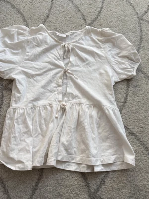 Vit blus med knyt från Gina Tricot - Söt vit blus från Gina Tricot med puffiga korta ärmar och två knyt framtill. Blusen har en avslappnad passform och volang nedtill. Tillverkad i mjukt bomullstyg, perfekt för en trendig och bekväm look. Passar XS/S.