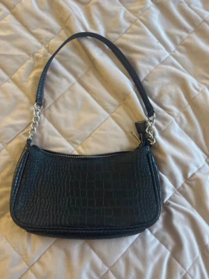 Svart axelväska i krokopräglat skinn - Trendig svart handväska i krokopräglat konstläder med kort axelrem och silverfärgade kedjedetaljer. Väskan har en stilren, avlång form och stängs med dragkedja upptill. Perfekt accessoar för att lyfta din outfit.