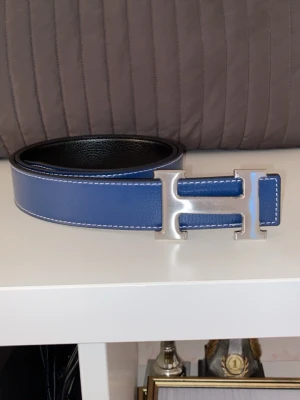 Blått bälte med H-spänne - Snyggt blått bälte med ikoniskt H-spänne i silverfärg. Bältet har vita sömmar och är gjort i slätt skinn. Perfekt accessoar för att lyfta din outfit med en exklusiv touch.