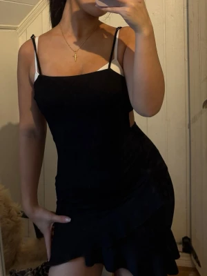 Svart kort klänning med knyt i ryggen - Säljer en svart kort klänning med smala axelband och snygg knytning i ryggen. Klänningen har en enkel, figurnära passform och volangdetalj nedtill. 