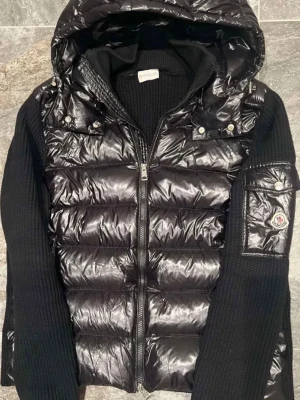 Svart pufferjacka från Moncler - Snygg svart pufferjacka från Moncler med glansig quiltad front och huva. Jackan har stickade svarta ärmar och detaljerad Moncler-logga på ena ärmen. Dragkedja framtill och fickor med tryckknappar. Perfekt för kalla vinterdagar.