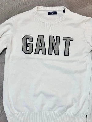 Vit stickad tröja från GANT - Säljer en vit stickad tröja från GANT med stora svarta bokstäver på bröstet. Tröjan har rund hals och ribbstickade muddar vid hals, ärmslut och nederkant. Perfekt för dig som gillar klassisk och stilren design.