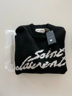 Svart stickad tröja Saint Laurent - Svart stickad tröja från Saint Laurent med vit logotyp i stor, handskriven stil över bröstet. Tröjan har rund halsringning och ribbstickade muddar. Perfekt för dig som gillar exklusiva och trendiga plagg.