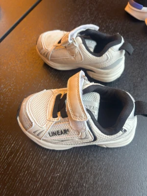 Vita sneakers från Linear med kardborre - Snygga vita sneakers från Linear med svart detalj och kardborreband. Skorna har meshpaneler för extra andningsförmåga och en sportig look. Perfekta för dig som gillar stilrena och bekväma sneakers med enkel påtagning.