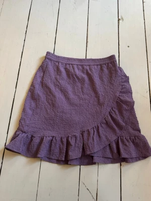 Lila volangkjol med struktur - Kort lila kjol med volangkant och omlott-look. Kjolen har en snygg struktur i tyget som ger extra detalj och är perfekt för dig som gillar en lekfull och trendig stil. Resår i midjan för skön passform.