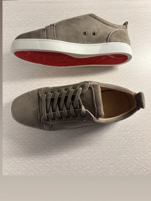 Gråa sneakers med röd sula - Snygga sneakers i grå mocka med vita sulor och röd undersida. Skorna har rund tå, snörning framtill och diskreta ventilationshål på sidan. Insidan är ljusbrun och materialet känns lyxigt. Perfekta för dig som gillar stilrena detaljer.