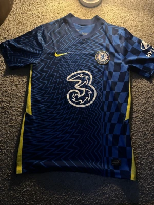 Chelsea FC matchtröja blå Nike - Chelsea FC matchtröja i blått med zigzag- och rutmönster, gula detaljer och klubbmärke på bröstet. Tillverkad i lätt och ventilerande material, perfekt för fotboll eller träning. Stort vitt sponsorsmärke på framsidan och Nike-logga i gult.