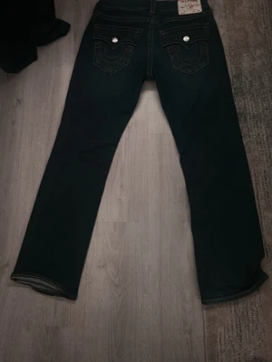 True Religion mörkblå bootcut jeans - Säljer ett par mörkblå jeans från True Religion med bootcut passform. Jeansen har ikoniska kontrastsömmar, stora fickor bak med lock och silverfärgade knappar. Klassisk patch med logga bak i midjan. Perfekta för en trendig och avslappnad stil.