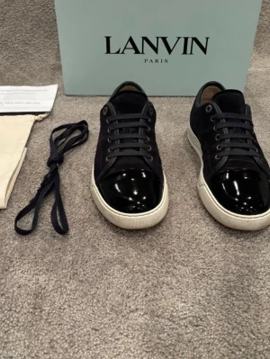 Mörkblå Lanvin DBB1 Cap Toe uk 6 - Snygga mörkblå sneakers från Lanvin i modellen DBB1 Cap Toe. Skorna har en unik lackad tå, mörkblå mocka överdel och vit sula. Materialmix av mocka och läder ger en lyxig känsla. Perfekta för dig som vill sticka ut med stilrena detaljer. De är i mycket bra skick. Box, dustbag och extra skosnören tillkommer vid köp. Hör av er vid frågor och funderingar!