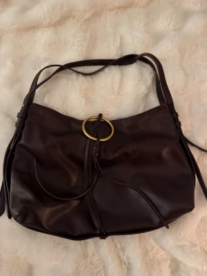 Mörkbrun axelväska med guldring - Snygg vinröd axelväska i mjukt läder med dekorativ guldfärgad ring framtill och långa remmar. Väskan har en avslappnad, slouchy form och detaljer i form av knutar och hängande läderband.