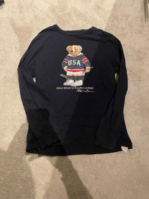 Mörkblå polo Ralph lauren tröja M - Mörkblå långärmad tröja från Ralph Lauren med Polo Bear på bröstet, iklädd hockeyoutfit med USA-tryck. Tröjan har rund hals och är gjord i mjuk bomull. Perfekt för dig som gillar sportiga och ikoniska plagg.